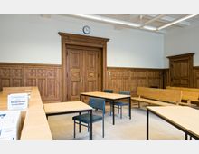 Ein Sitzungsraum mit Holzwandvertäfelungen und Holztüren im klassischen Stil. Der Raum ist mit einfachen Holztischen, Stühlen mit Stoffpolsterung und Holzbänken ausgestattet, eine Wanduhr hängt über einer Tür.