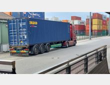 Ein roter LKW mit Auflieger transportiert einen blauen Frachtcontainer auf einer Straße. Im Hintergrund sind gestapelte farbige Container und ein Zaun zu sehen, die auf ein Hafengelände oder eine Lagerung an einem Containerterminal hinweisen.