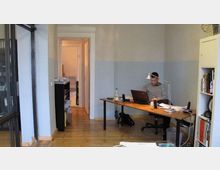 Ein schlichtes Büro mit Holzboden und hellgrauen Wänden; im Vordergrund ein Schreibtisch mit einem Laptop und Arbeitsmaterialien, an dem eine Person sitzt. Rechts steht ein Regal mit Büchern, und im Hintergrund führt ein Durchgang zu einem weiteren Raum.