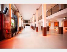 Ein großer, industrieller Innenraum mit hohen Decken, weißen Wänden und braun gestrichenen Säulen. Der Raum verfügt über große Fenster mit Pflanzen an den Fensterbänken, sichtbare Rohrleitungen und Geländer im Obergeschoss, die eine erhöhte Ebene andeuten.