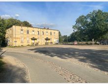 Ein historisches, zweistöckiges Gebäude mit verblasster Fassade und hohen Fenstern steht an einem Platz mit gepflasterten Wegen. Im Hintergrund sind Bäume und einzelne Schilder sowie Bushaltestellen zu sehen.