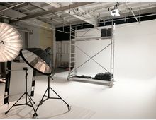 Fotostudio mit weißem Hintergrund, großen Softboxen auf Stativen und einem Gerüst im Zentrum. Der Raum hat eine industrielle Decke mit sichtbaren Balken und Beleuchtungseinrichtungen.