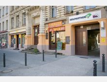 Straßenszene mit mehreren Geschäftseingängen in einem historischen Wohn- und Geschäftsgebäude mit verzierten Fassaden. Zu sehen sind ein Imbiss, ein Geschäft namens "Seilpartner" und eine Apotheke, sowie eine offene Durchfahrt zu einem Innenhof.