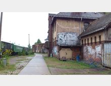 Ehemaliges Bahngelände mit einem alten, mit Graffiti bedeckten Backstein-Gebäude und einer stillgelegten, grünen Eisenbahn auf einem Gleis. Der Boden besteht aus Kopfsteinpflaster und Beton, umgeben von vereinzelten Pflanzen und Büschen.