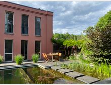 Rückansicht eines modernen, zweigeschossigen Hauses mit rosafarbener Fassade und hohen Fenstern, davor eine Terrasse mit Holztisch und Stühlen. Im Vordergrund liegt ein naturnaher Gartenteich mit Seerosen und Trittplatten, umgeben von dichter Bepflanzung und Sträuchern.