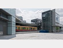 Moderner Bahnhof mit einer gelben und roten S-Bahn auf einem erhöhten Bahngleis. Rechts im Bild ein gläsernes Gebäude mit der Beschriftung "DB Parkhaus Süd" und einer großen Uhr auf der Fassade.
