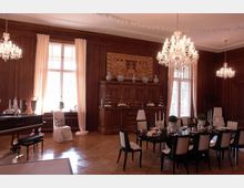 Elegantes Speisezimmer mit holzvertäfelten Wänden, Kronleuchtern und großen Fenstern mit weißen Vorhängen. Ein reich gedeckter Esstisch und ein dekorativer Holzschrank mit verzierten Reliefs stehen im Mittelpunkt.