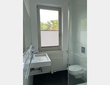 Modernes Badezimmer mit weißer Ausstattung, bestehend aus einem Wandwaschbecken, einem Hänge-WC und einer gläsernen Duschkabine. Ein großes Fenster mit halb geschlossenem Plissee lässt Tageslicht herein, während der Boden mit dunklen Fliesen gestaltet ist.