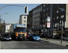 Straßenszene in einer Stadt mit mehrstöckigen Wohn- und Geschäftsgebäuden, darunter eine Apotheke. Im Vordergrund fahren ein großer LKW und mehrere Autos, während eine Uhr und eine Werbesäule am Straßenrand sichtbar sind.