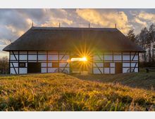 Fachwerkgebäude mit Strohdach in einer ländlichen Umgebung, durch dessen offene Toröffnung die tiefstehende Sonne strahlt; davor eine Wiese im Abendlicht, im Hintergrund einzelne Bäume und ein wolkiger Himmel.