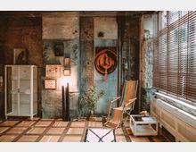 Industrieller Wohnraum mit Sichtziegelwänden und metallischen Rohren, eingerichtet mit einem weißen Vitrinenschrank, einem modernen Lampenständer, zwei gestreiften Liegestühlen und einer Pflanze. Große Fenster mit Jalousien lassen Tageslicht in den Raum, während der Holzboden das rustikale Ambiente ergänzt.