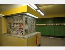 Kleiner Kiosk mit Zeitschriften hinter einem vergitterten Schaufenster in einer U-Bahn-Station mit grünen Wandpaneelen und der Beschilderung „Eisenacher Straße“.