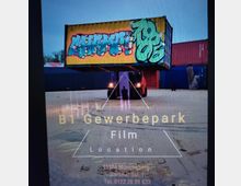 Ein großer gelber Container mit Graffiti-Beschriftung wird von einem Gabelstapler auf einem offenen Industriegelände bewegt. Im Hintergrund sind weitere Container sowie eine asphaltierte Fläche zu sehen.