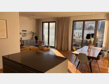 Modernes Wohnzimmer mit Essbereich und großer Fensterfront, die einen Blick auf eine Stadtlandschaft bietet. Der Raum hat Holzfußboden, eine offene Küche mit dunkler Arbeitsfläche, einen Esstisch mit Stühlen und eine gemütliche Sitzecke mit Pflanzen und Dekoration.