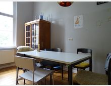 Helles Esszimmer mit einem weißen Tisch und verschiedenen Stühlen, teils mit Sitzauflagen. Links steht ein Holzvitrinenschrank, rechts an der Wand ist Kinderkunst mit Sternen und einem Hasen angebracht.