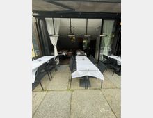 Außenbereich eines Restaurants mit gedeckten Tischen und weißen Tischdecken, umgeben von schwarzen Stühlen. Im Hintergrund ist der Innenraum mit weiteren gedeckten Tischen und heller Beleuchtung zu sehen, eingerahmt von weißen Vorhängen.