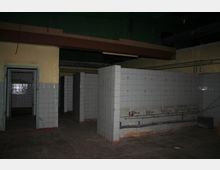 Ein leerer, alter Raum mit gefliesten Wänden und wenigen Trennwänden. An einer Wand befinden sich mehrere Wasseranschlüsse mit sichtbaren Rohren, die auf mögliche frühere Nutzung, wie etwa eine Waschanlage, hinweisen.