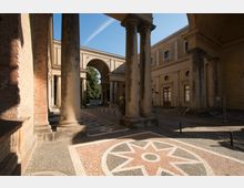 Historischer Innenhof mit Arkaden aus gelbem Backstein, mehreren Rundbögen und Säulen. Der Boden ist mit detaillierten Mosaikmustern in hellen und dunklen Steinen gestaltet, umgeben von schattigen und sonnigen Bereichen.