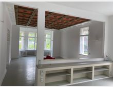 Helles Zimmer mit großen, weißen Sprossenfenstern und einer verzierten, rot-braunen Kassettendecke. Der Raum verfügt über weiße Wände, einen hellgrauen Boden und eingebaute Regale unterhalb einer Durchreiche.