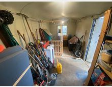 Ein schmaler Abstellraum mit Betonboden, gefüllt mit Gartengeräten wie Rechen und Schaufeln, sowie Regalen mit verschiedenen Gegenständen. Im Hintergrund befindet sich ein Holzgitter, das den Raum teilt, und eine Tür führt nach draußen.