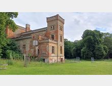 Eine alte, teilweise verfallene Villa mit Backsteinmauern und einem rechteckigen Turm auf der linken Seite, umgeben von einer Wiese und großen Bäumen im Hintergrund. Das Gebäude zeigt Spuren von Erosion und eingestürzten Elementen, während der Hof durch einen niedrigen Zaun abgetrennt ist.