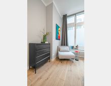 Heller Wohnraum mit hohen Decken, großem Fenster mit halbhoher Milchglasfolie und grauen Vorhängen sowie hellem Holzboden. Neben einer schwarzen Kommode mit Vase stehen eine gepolsterte Liege und ein kleiner Beistelltisch vor dem Fenster, an der Wand hängt ein farbiges Bild.