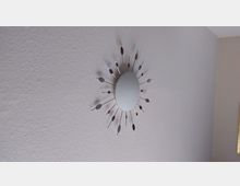Wand mit einer dekorativen, runden Wanduhr in silberner Farbe, die strahlenförmig mit metallischen Elementen gestaltet ist. Der Hintergrund besteht aus einer einfarbigen, beige gestrichenen Wand.