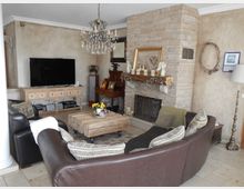 Wohnzimmer mit einer halbrunden braunen Ledercouch, einem rustikalen Holztisch in der Mitte und einem Kamin mit Steinverkleidung. Im Hintergrund befinden sich ein großer Flachbildfernseher auf einer Holzkommode, ein Kronleuchter und dekorative Elemente wie Bilderrahmen, Figuren und ein Wandkranz.