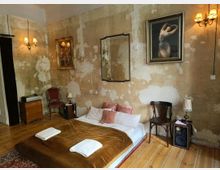 Schlafzimmer mit rustikalem Charakter, gekennzeichnet durch abgeblätterte Wände, einen niedrigen Futon mit braunem Überwurf und weißen Handtüchern, Holzboden und antiken Möbeln. An den Wänden hängen Gemälde und ein Spiegel, beleuchtet von Wandleuchten mit warmem Licht.