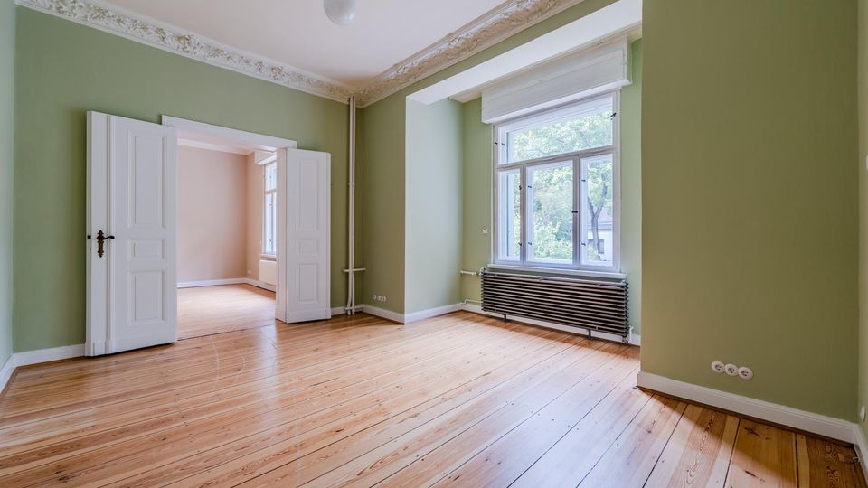 Leerer Altbau-Wohnraum mit hellen Holzdielen, grünen Wänden und dekorativem Stuck an der Decke. Große Fensterfront mit Heizkörper darunter sowie eine doppelflügelige Tür, die in einen weiteren Raum führt.