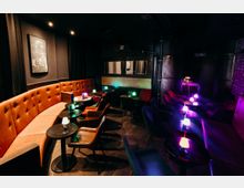 Innenraum einer dunklen Bar-Lounge mit gepolsterter, orangefarbener Sitzbank entlang der Wand, kleinen runden Tischen und Sesseln. Farbige Tischlampen und gedämpfte Deckenbeleuchtung setzen Akzente, im Hintergrund sind eine abgetrennte Nische und ein Notausgangsschild zu sehen.