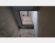 Blick nach oben in einem vertikalen Schacht mit betonierten Wänden. Ein Geländer in grüner Farbe ist sichtbar, darüber eine Öffnung mit Licht, und ein Draht oder Seil hängt durch den Schacht.