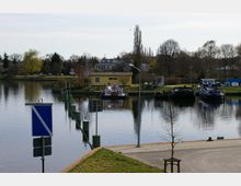 Uferbereich eines Kanals mit befestigter Promenade im Vordergrund. Im Wasser liegen mehrere kleine Schiffe und ein gelbes Gebäude steht am gegenüberliegenden Ufer, umgeben von Bäumen und Sträuchern.