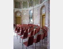 Prunkvoller barocker Saal mit verzierten Wänden, Deckengemälden und einem großen Spiegel in der Mitte, eingerichtet mit roten Stühlen, die in Reihen im Halbkreis aufgestellt sind. Holztüren und ein heller Teppich komplettieren den Raum.