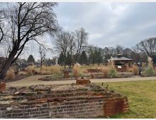 Ein Park mit einer Rasenfläche, geschwungenen Wegen und dekorativen Ziergräsern. Im Hintergrund steht ein hölzerner Pavillon, während im Vordergrund eine alte, teilweise verfallene Backsteinmauer zu sehen ist.