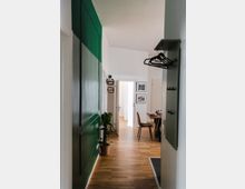 Schmaler Flur mit Holzboden, links eine grüne Wand mit Pflanzen und rechts eine Garderobe mit Kleiderbügeln. Im Hintergrund ein Essbereich mit Tisch, Stühlen und dekorativem Geschirr, umgeben von weißen Wänden und Bildern an der Wand.