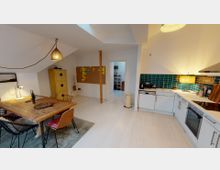 Offene Wohnküche mit Holzboden und Dachschrägen. Links steht ein Esstisch mit gemischten Stühlen und einer Hängelampe, rechts eine moderne Einbauküche mit weißer Front und grünen Fliesen als Rückwand; im Hintergrund ein Durchgang zu einem angrenzenden Raum.