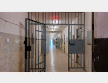 Ein schmaler Gefängnisflur mit offen stehenden Metallgittern im Vordergrund. Entlang der Wände befinden sich Zellentüren aus Metall, und die abblätternde Wandfarbe zeigt Abnutzungsspuren; eine rote Warnleuchte ist an der Decke sichtbar.