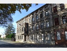 Fassade eines alten, mehrstöckigen Wohngebäudes mit detaillierten Verzierungen und sichtbaren altersbedingten Schäden. Im Vordergrund verläuft eine leere Straße, während auf der linken Seite Bäume und vereinzelte kleinere Gebäude zu sehen sind.