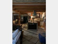 Rustikaler Wohn- und Küchenbereich mit freiliegenden Holzbalken, einem Küchenblock in der Mitte und gemusterten Fliesenböden. Im Hintergrund sind ein Holzschrank, eine Wendeltreppe und Wände mit natürlichen Stein- und Putzakzenten zu sehen.