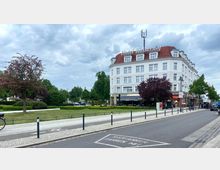 Straßenszene mit einem mehrstöckigen Gebäude im klassischen Stil, auf dessen Dach „Hotel Kaiserhof“ steht. Im Vordergrund Grünflächen mit Bäumen und Gehwegen, daneben eine Straße mit markiertem Radweg und parkenden Autos.