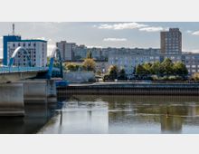 Stadtansicht mit einer blauen Bogenbrücke im Vordergrund über einem Fluss. Im Hintergrund stehen Wohngebäude und Hochhäuser, umgeben von Bäumen und einer Uferpromenade.