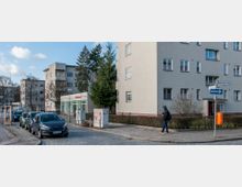 Straßenszene in einer Wohngegend mit mehrstöckigen, hellen Wohngebäuden im Bauhaus-Stil, parkenden Autos am Straßenrand und einem kleinen Geschäft mit der Aufschrift „Bäckerei“. Ein Fußgänger läuft auf dem Gehweg, daneben Straßenschilder und ein oranger Briefkasten.