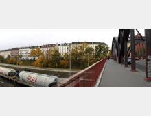 Eine Brücke mit rotem Geländer und stählernen Bögen führt über eine Bahntrasse, auf der ein Güterzug mit Kesseltankwagen steht. Im Hintergrund sind mehrstöckige Wohnhäuser aus der Gründerzeit sowie herbstlich gefärbte Bäume zu sehen.