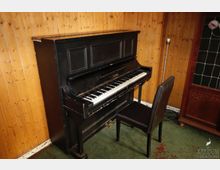 Schwarzes Klavier vor einer holzvertäfelten Wand mit einem gepolsterten schwarzen Stuhl davor. Im Hintergrund sind ein Stehlampe und ein Holzschrank mit Glaseinsatz sichtbar.