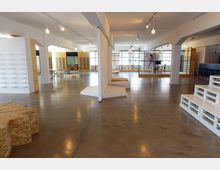 Großer, offener Raum mit modernem Industriestil, Betonboden, weißen Wänden und deckenhohen Fenstern, die viel Licht einlassen. Der Raum enthält modulare Möbel aus Holz und Metall sowie Regale und Podeste, teilweise aus weißen Paletten gefertigt.