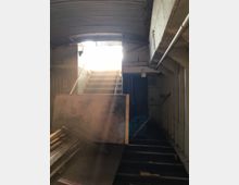 Ein verlassener, dunkler Treppenaufgang in einem industriellen Raum mit Betonwänden und einem gewölbten Metalldach; Sonnenlicht scheint durch den oberen Ausgang hinein, während Holzlatten und Materialien den unteren Bereich teilweise blockieren.