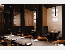 Elegantes Restaurant mit moderner Einrichtung, dunklen Sitznischen und hölzernen Stühlen. Die Beleuchtung ist warm und dezent, mit weißen, runden Hängelampen und dekorativen Elementen wie einem goldfarbenen Zweig in der Tischmitte.