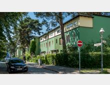 Straßenszene in einer Wohngegend mit grün gestrichenen Reihenhäusern im Bauhaus-Stil, umgeben von Bäumen und Sträuchern. Am Straßenrand stehen geparkte Autos, und Straßenschilder markieren den "Treibjagdweg" und "Am Heidewinkel".