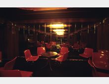 Raum mit dunkler Beleuchtung, roten Kunststoffstühlen und runden Tischen, umgeben von Spiegelwänden, die einen großen, leuchtenden Kronleuchter reflektieren. Die Atmosphäre wirkt intim, mit dunklen Holz- und Vorhang-Elementen.